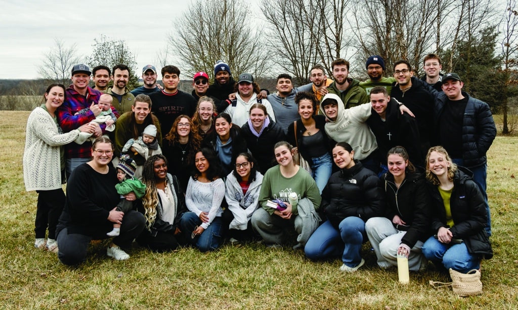 A large group of young adults and a few small children pose together outdoors on a cloudy day, smiling warmly at the camera. They are bundled up in winter and fall clothing, standing on a grassy field with bare trees in the background.